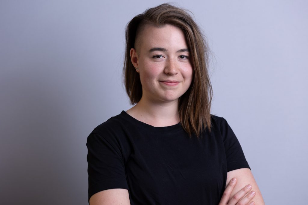 Zu sehen ist Kathi Broermann. Sie lächelt in die Kamera. Sie hat schulterlange Haare auf einer Seite. Auf der anderen Seite trägt sie kurz rasierte Haare. Sie trägt ein schwarzes Shirt. Am unteren Bildrand sind die verschränkten Arme angedeutet.